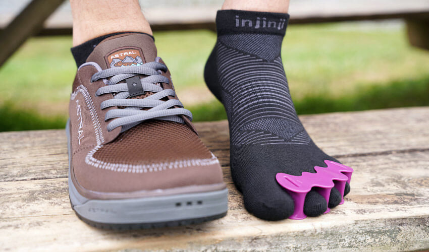 A set of feet resting on a wooden picnic table seat, one foot wearing a Correct Toes Plum toe spacer and Injinji toe sock, the other foot wearing an Astral shoe