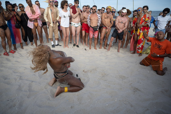 Keoui dances on her knees on the beach, blonde hair flipping back, with an audience encircling her