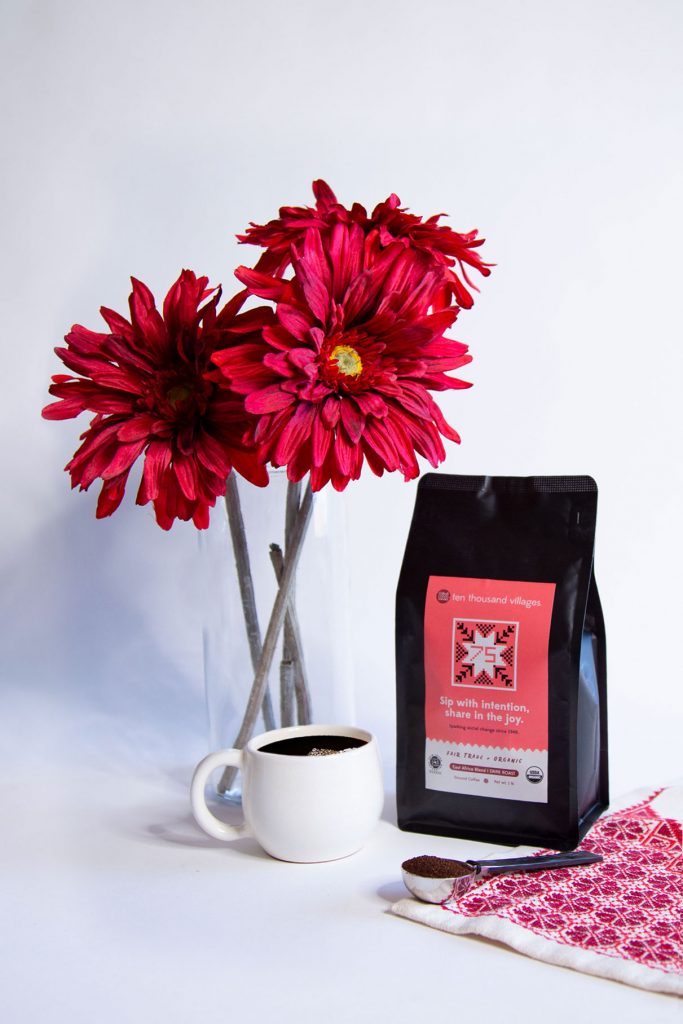Three large red flowers stand in a vase behind a white mug of fair trade coffee. Next to these things are a bag of Ten Thousand Villages' 75th Anniversary coffee, a scoop of ground coffee, and a red and white embroidery.