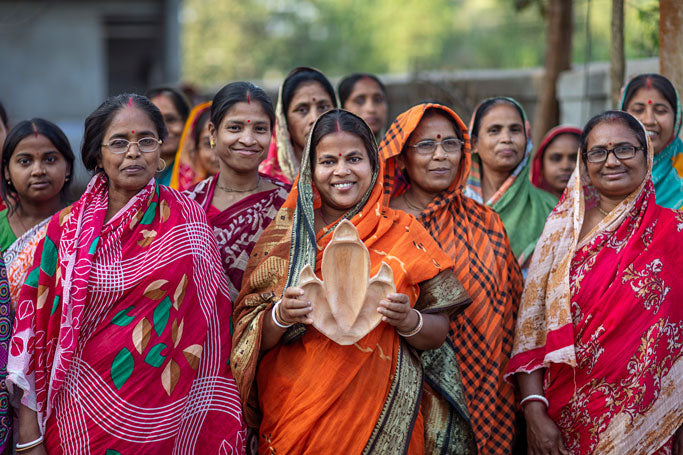 A group of the women artisans in India who handcrafted much of the fair trade movie merchandise collection by Jurassic World x Accompany is shown standing together in colorful sari garments. The woman front and center smiles. She has a bindi on her forehead and holds up a Tyrannosaurus Rex Plate. 