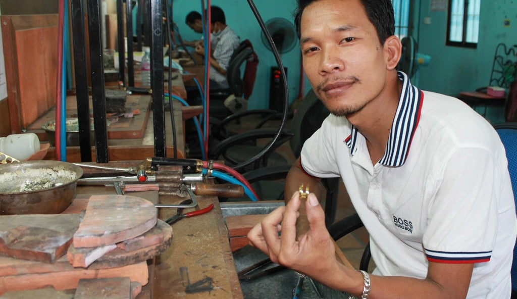 Rajana artisan, Heng Sopheanith is pictured in a workshop, seated at a workstation, holding up a bombshell ring he has fashioned from the recycled bombshells. 