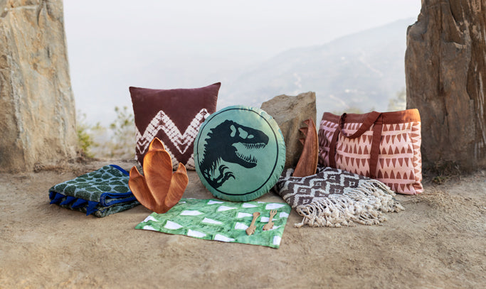 Pieces from the Jurassic World x Accompany movie merchandise collection sit on a hillside in Nepal where many were handcrafted. The T. Rex Born To Be Wild pillow is front and center with other items surrounding it. 