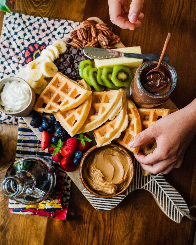 Image is of a brunch board on the Minimalist Serving Board from Ten Thousand Villages. A hand reaches for waffles that are surrounded by berries, kiwi slices, peanut butter, cocoa spread, banana slices, chocolate chips and butter with the Butter Together Knife. 