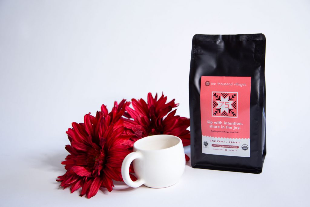 Bag of Ten Thousand Villages' 75th Anniversary coffee sits with a white mug and red flowers on a white background. Bag label says 
