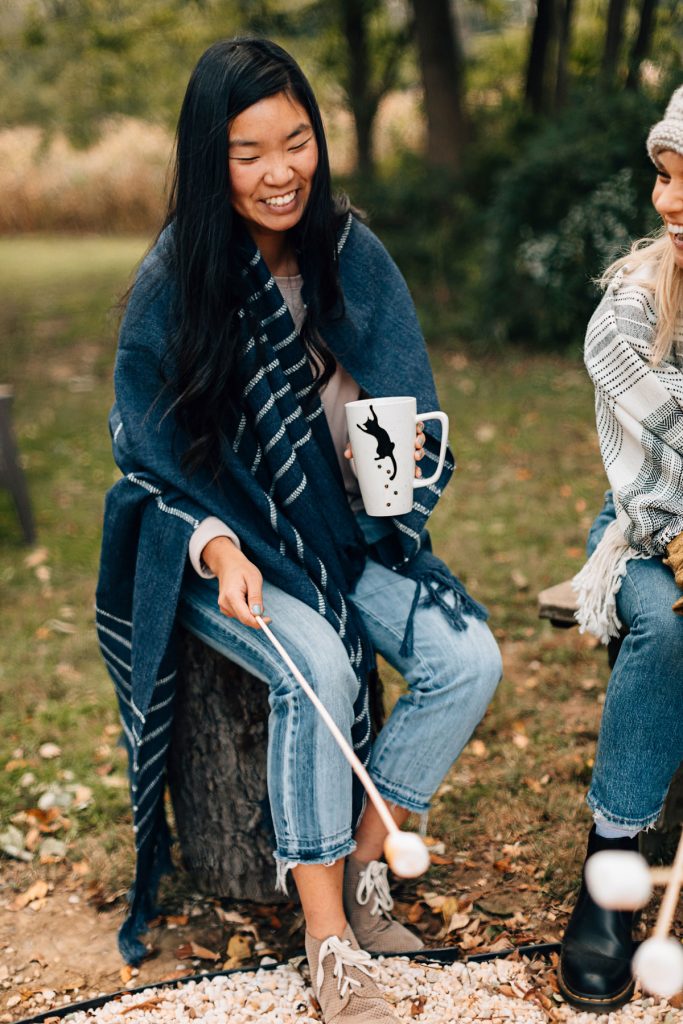 How to make s'mores better. Image shows an Asian woman with long dark hair wrapped in a handwoven blanket. She's holding a handcrafted mug with the silhouette of black cat on it. She's seated next to another woman who is just out of frame and both are roasting marshmallows over a campfire. 