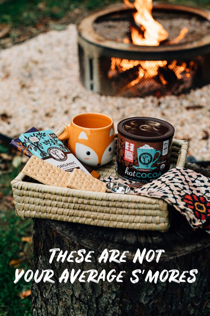 How to make s'mores better. Image shows a handcrafted mug in the shape of a fox face nestled in a handwoven grass basket with Equal Exchange hot cocoa, graham crackers, a recycled sari napkin and Equal Exchange chocolate bars. In the background is a lit campfire. 