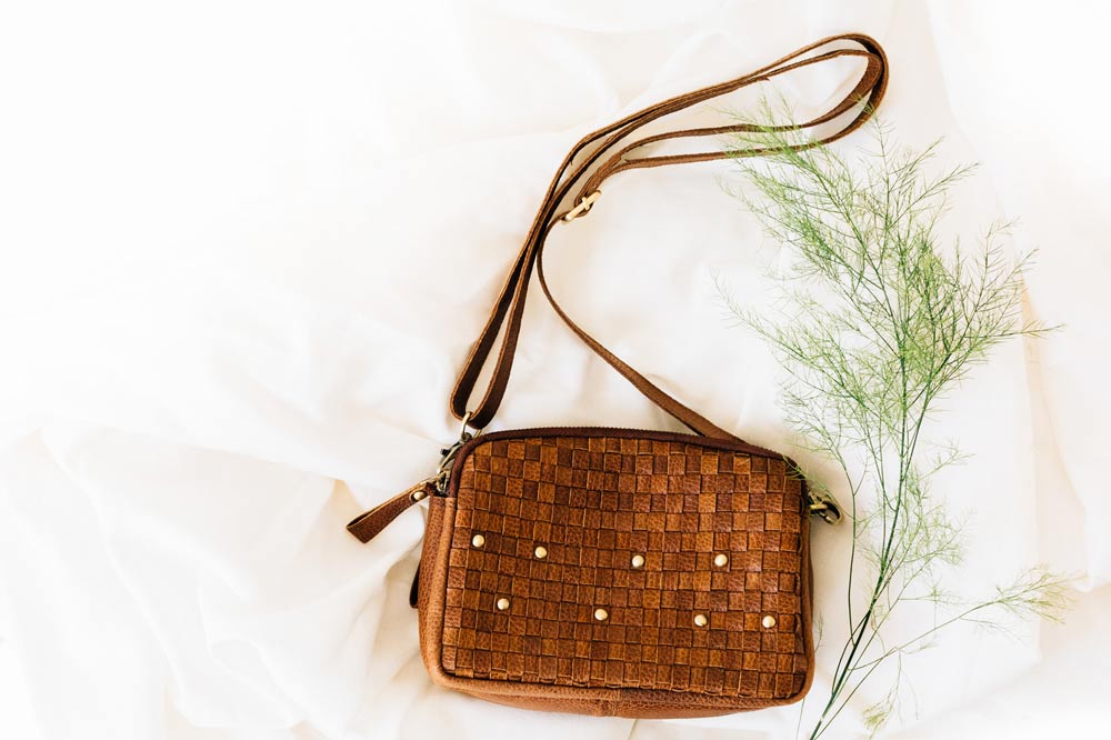 Summer 2021 Fashion Trend: The Eco-Leather Woven Purse, a brown leather woven purse with brass accents lies on linens with a sprig of greenery.