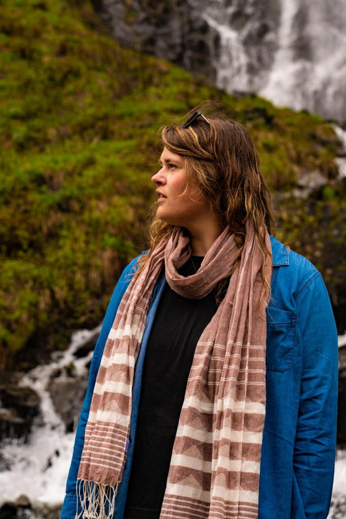 Model looks off in the distance wearing the Jamdani Weave Scarf around her neck over a blue chambray shirt. Waterfalls crash behind her. 