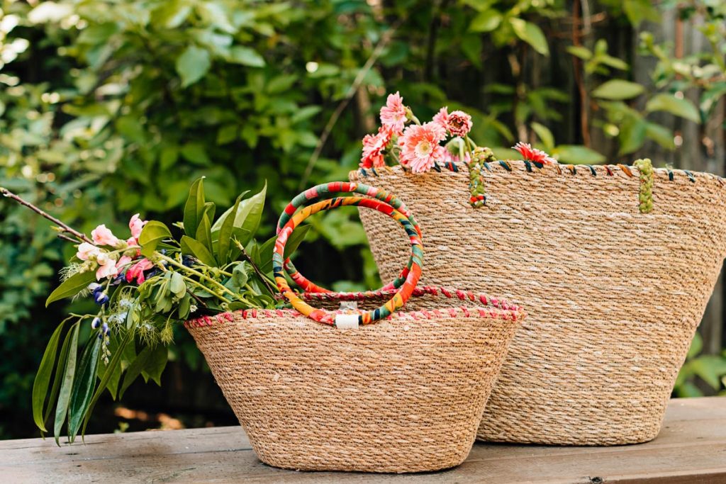 Trend-forward woven bags sit one in front of the other with fresh flowers in them. Handwoven bags with natural grasses and upcycled sari. 