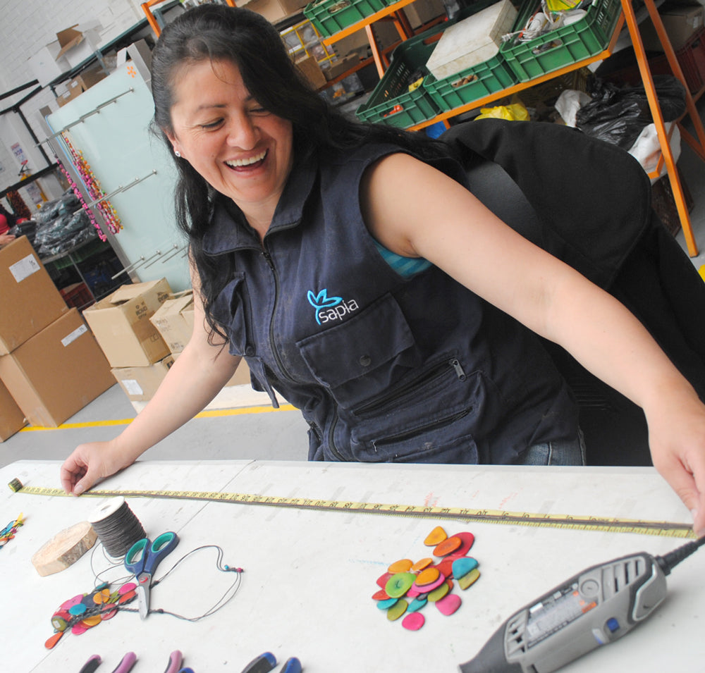 Columbian artisan measures cord for a sustainable jewelry piece she is making out of tagua. Colorful tagua pieces lay on the table in front of her and she is laughing. 