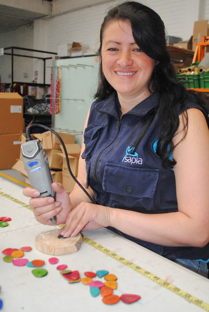Claudia Zambrano, artisan, is wearing a Sapia vest, working in a workshop at a table. She is using an electric drill on bright pieces of tagua. She is smiling at the camera. 