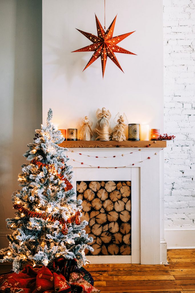 How to stye a holiday mantle in traditional christmas colors. Photo shows a large red paper lantern above the mantle, handmade sinamay and burlap angels anchor the mantle and a flocked evergreen tree is in front with red garland. 