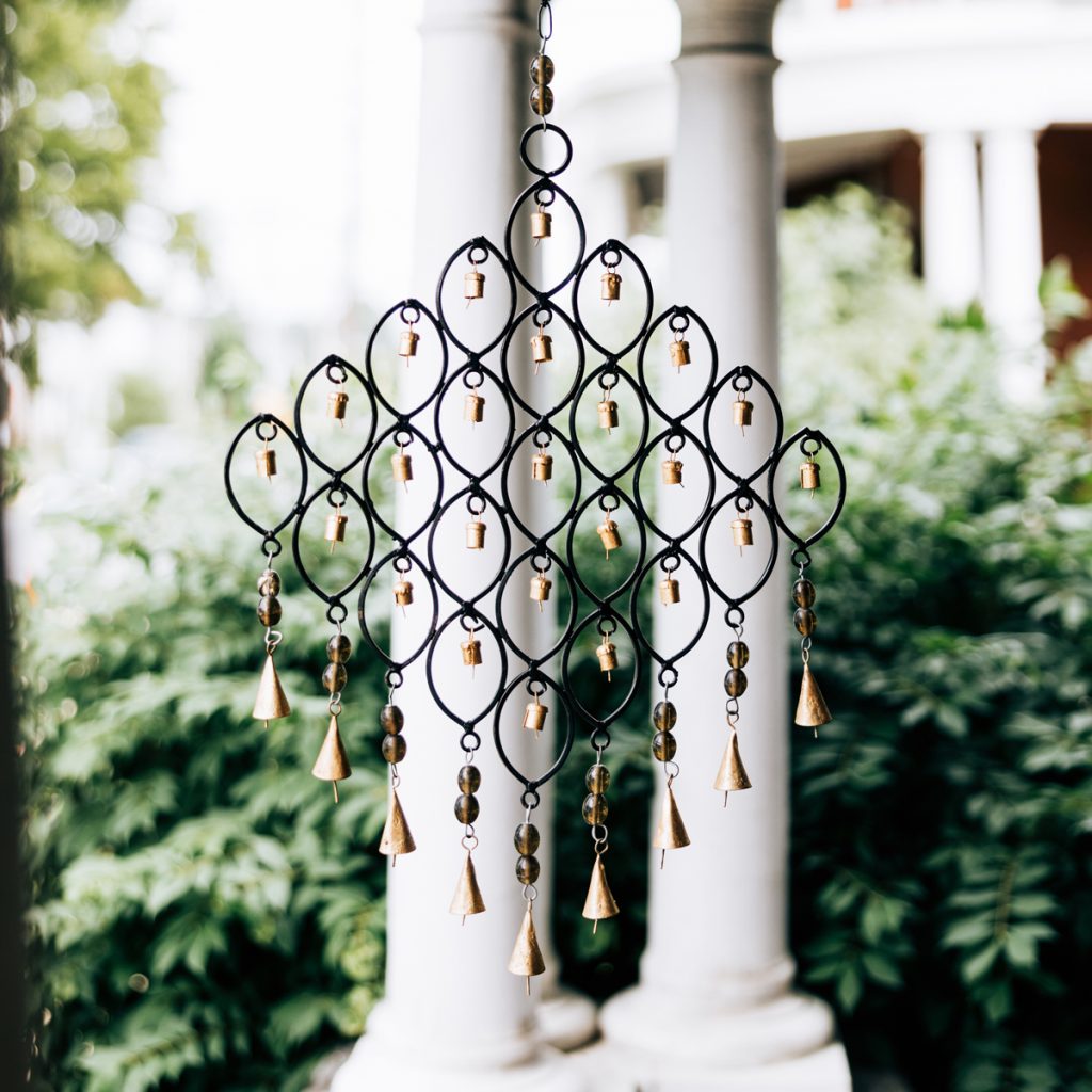 metal bell sculpture hanging on a porch with two striking white columns in the background with greens