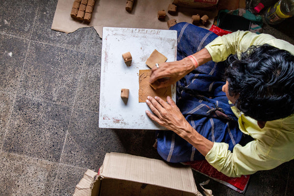 Image is taken from a bird's eye view. Features a person with black hair seated on a tile floor with a worktable in front of them. You see their hands working the natural matter that will become cone incense. They wear a light green shirt with a blue and white skirt or blanket over their lap. 