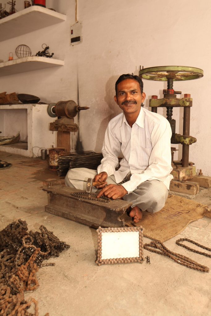 Artisan, Kamrul Hasan, is pictured seated on a mat in a workshop hammering bike chain. He is smiling at the camera and has a product sample of a bike chain picture frame in front of him that he created. 