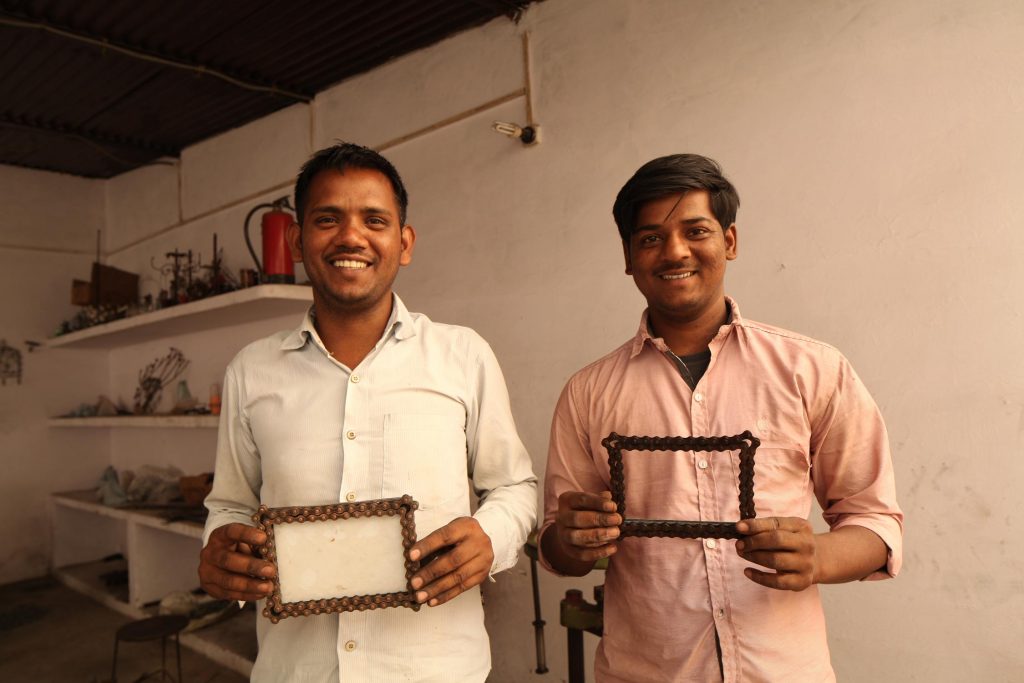 Two Noah's Ark artisans smile at the camera, each holding up a Bicycle Chain Picture Frame from our gifts for cyclists collection.