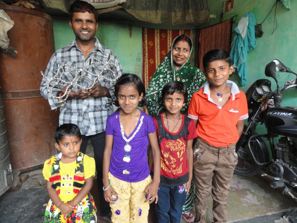 Artisan, Attar Singh and his family are pictured. He stands next to his wife with his four children in front of them. Attar Singh holds up a Bike Chain Wine Rack. There are sari textiles hanging behind them and a motorcycle parked to their right. 