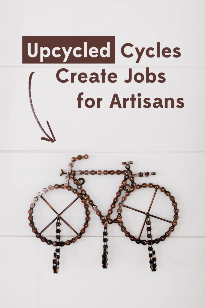 Upcycled Cycles Create Jobs for Artisans headline on a photo of a piece of upcycled bicycle chain art. The bicycle chain has been shaped into a bike with three hooks on it so it functions as hanging wall hooks. 