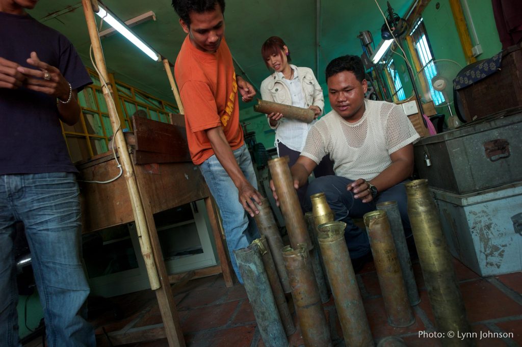 In a workshop in Cambodia, 4 members of the Rajana Association stack and inspect salvaged bomb shells. They are brass and 2-3 feet long. 