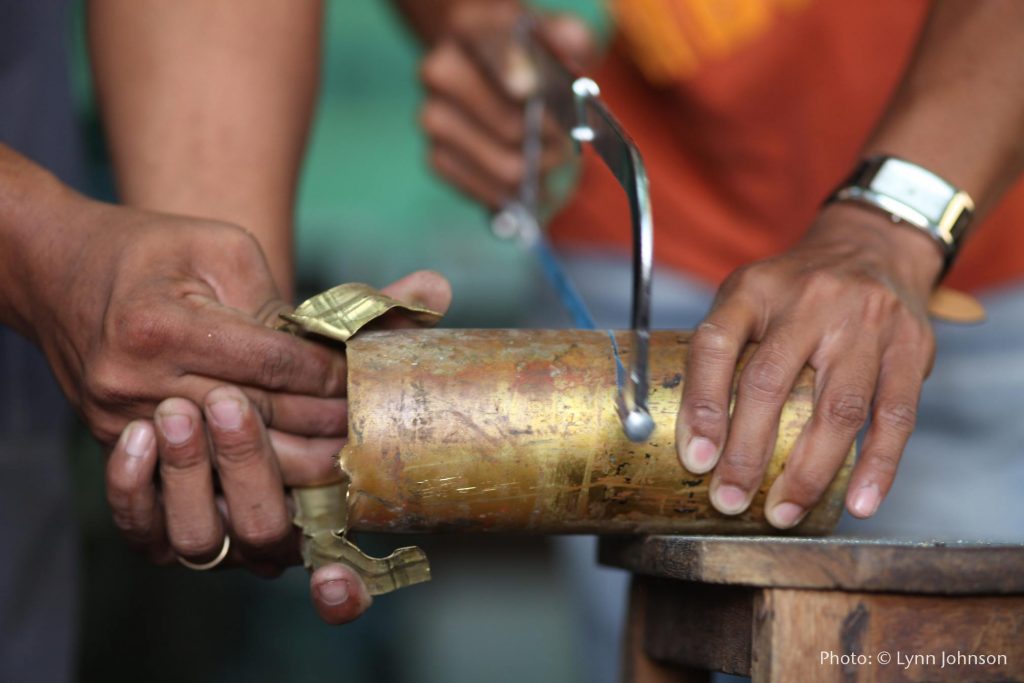 Two artisans work together to cut brass pieces off a bombshell to be melted down and turned into jewelry. Image shows one person's hands holding down the bombshell and sawing through it, while another set of hands peels pieces of brass metal away that have been cut. 
