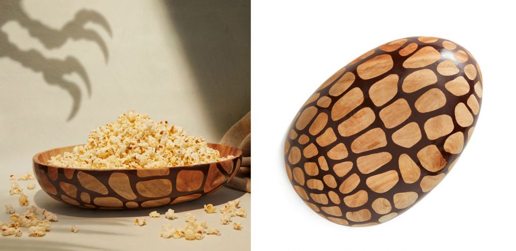 A split image of the Mega Feast Bowl from the Jurassic World x Accompany fair trade movie merchandise collection at Ten Thousand Villages. Shown full of popcorn with a shadow cast on the wall behind the bowl of raptor claws reaching for the popcorn. The right side of the image shows the bottom of the bowl, which features a dinosaur scale design. 