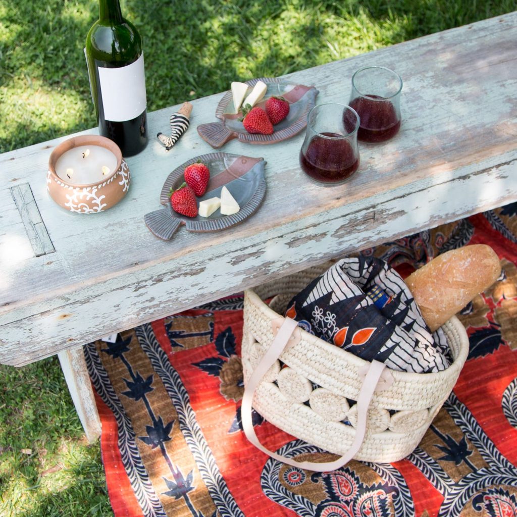 Sustainable picnic fare from Ten Thousand Villages is featured with a recycled sari blanket on the grass, the Essential Companion woven tote on top with another Recycled Sari Throw blanket inside. Picnic food and wine are laid out. 