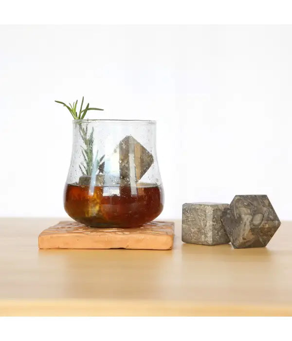 A cocktail glass sits on a stone coaster. The glass has a dark beverage with two Fossil Stone Drink Chillers and a sprig of rosemary. Two drink chillers sit on the table next to the glass as well. 