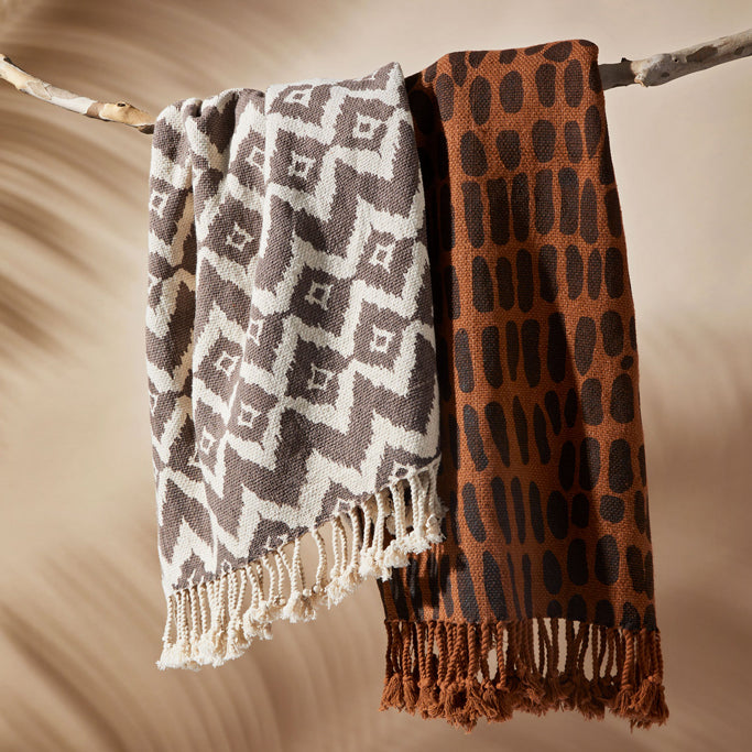 Two Keep It Wild blankets in two different color ways hang over a branch in front of a tan colored background with palm shadows on it. Blankets are from the Jurassic World x Accompany collection at Ten Thousand Villages. 