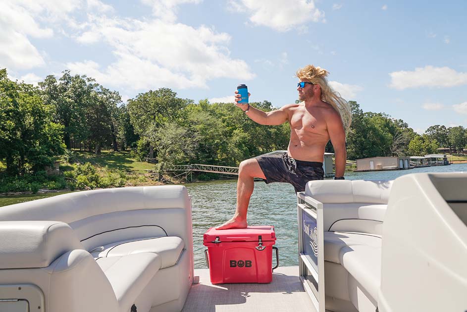 25QT / 23.6L vice red hard cooler on the deck of a boat with Chuck Beaver placing a foot on top and holding a drink in a can cooler.