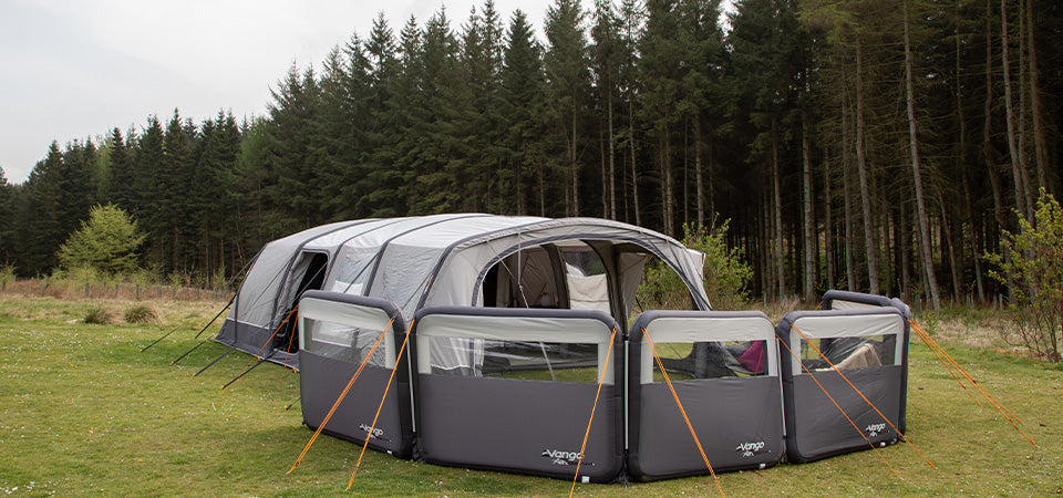 A large family tent pitched beside a forest.  In front of the tent is a premium Vango Modular Air Windbreak setup