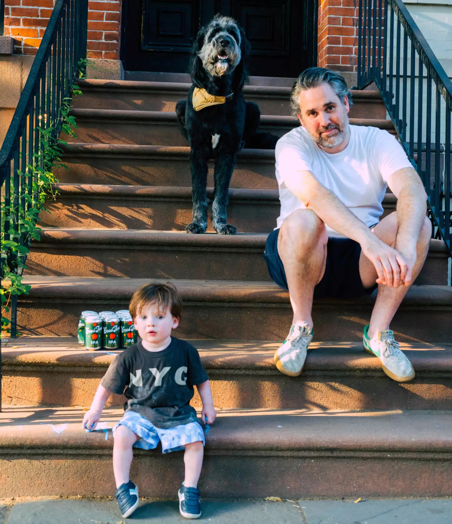 Michael 'Mikey' McFerran, his son, and his dog all sitting on a set of stairs. Also pictured is a 6 pack of Good Time IPA Non Alcoholic beers.