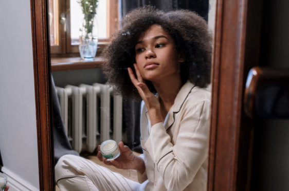 Une jeune femme est assise devant un miroir en pyjama, elle est en train de s’appliquer de la crème de soin anti-rides à l’acide hyaluronique sur le visage.