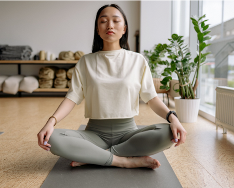 Une femme est assise sur un tapis de yoga et se tient en tailleur, les mais sur les genoux et les yeux fermés.