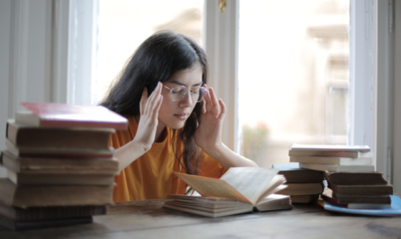 Une jeune étudiante se tient les temps dans un mouvement de fatigue intense. Devant elle se trouvent beaucoup de livres éparpillés et ouverts.