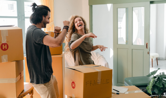 Un homme et une femme sont en train de danser dans leur salon avec beaucoup d’énergie. Ils rigolent en défaisant les cartons d’un déménagement.