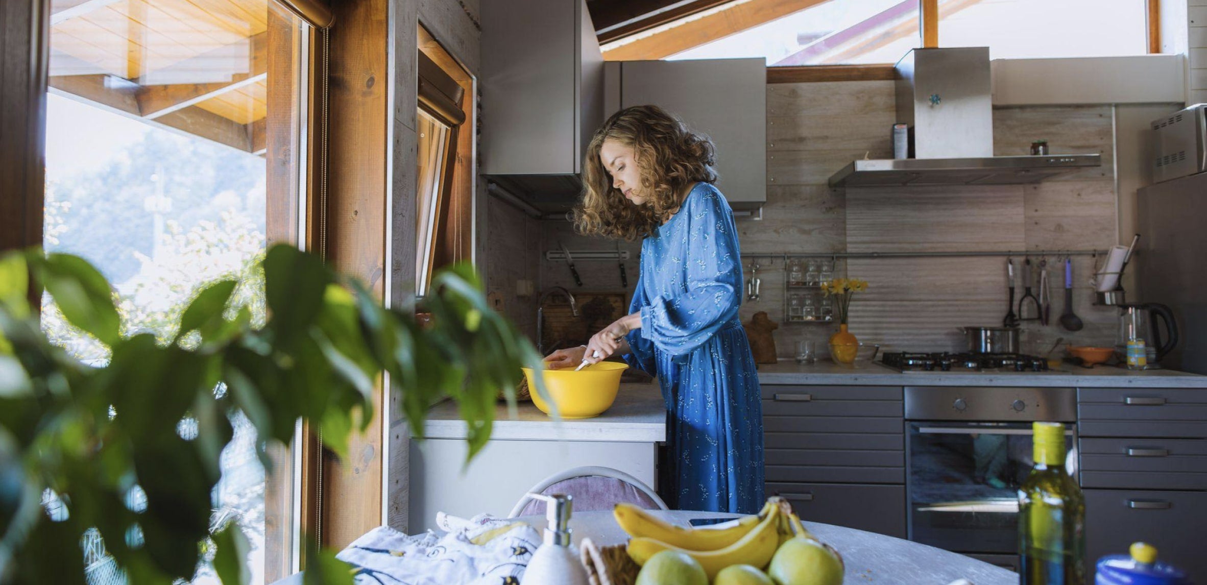 Une jeune femme est en train de cuisiner dans une cuisine ouverte et lumineuse. Elle se trouve au second plan et devant elle on peut voir une table sur laquelle se trouvent différents ingrédients comme des pommes, une banane, de l’huile d’olive et une plante.