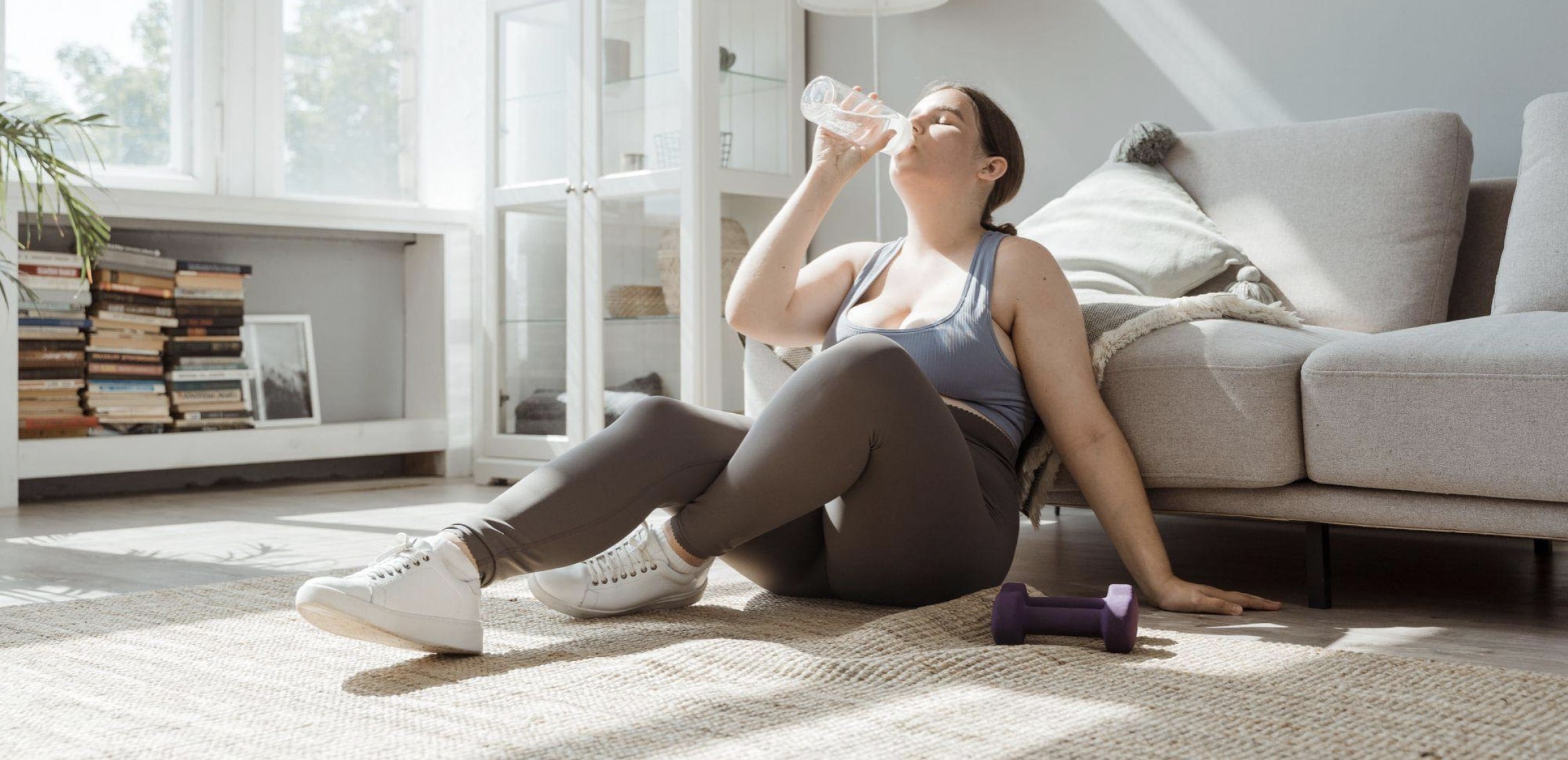 Une jeune femme est assise par terre, adossée à son canapé, après une séance de sport pour calmer son anxiété. Elle boit de l’eau et a une haltère pose à côté de sa main gauche.