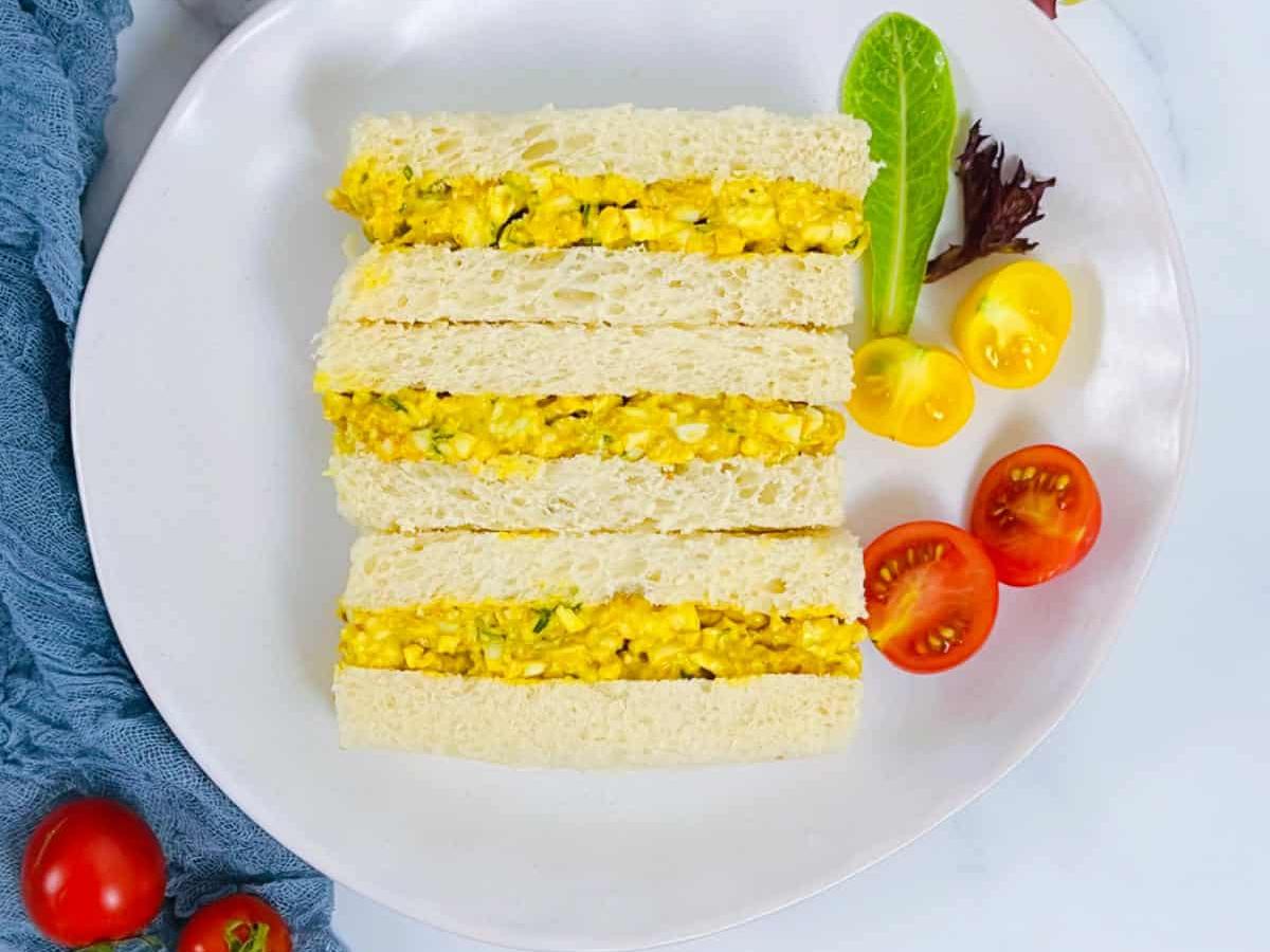 Curried egg sandwiches cut into neat rectangles, served on a white plate with cherry tomatoes and lettuce garnish
