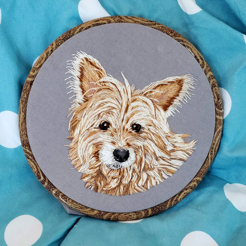 A hand embroidrred portrait of a terrier mix. His fur is orange and the fabric is grey
