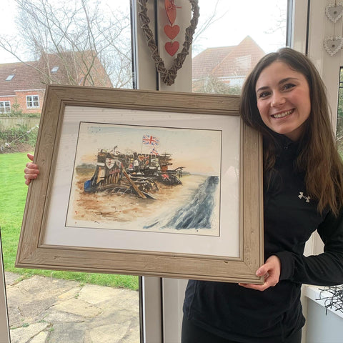 Cleethorpes artist, Eve Leoni Smith, holding a framed original watercolour painting of the Buck Beck Beach Bench on Cleethorpes Beach.
