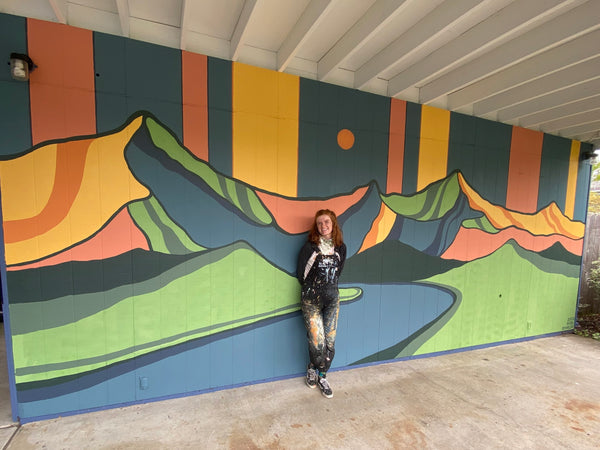 Nori leaning against a wall on which she has painted a colorful mural of mountains. 