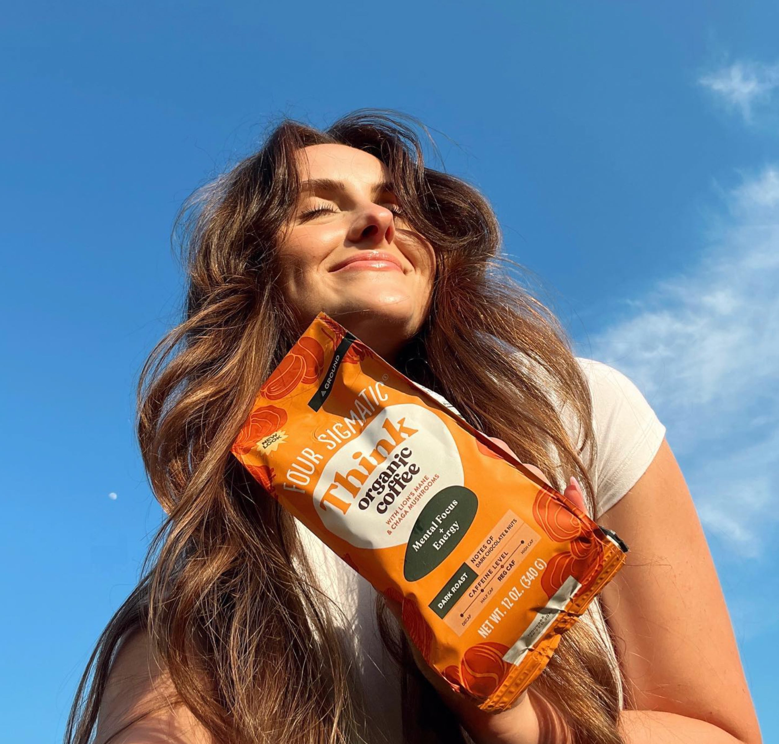 Woman smiling while holding a bag of Four Sigmatic Think Organic Coffee under a clear, blue sky.