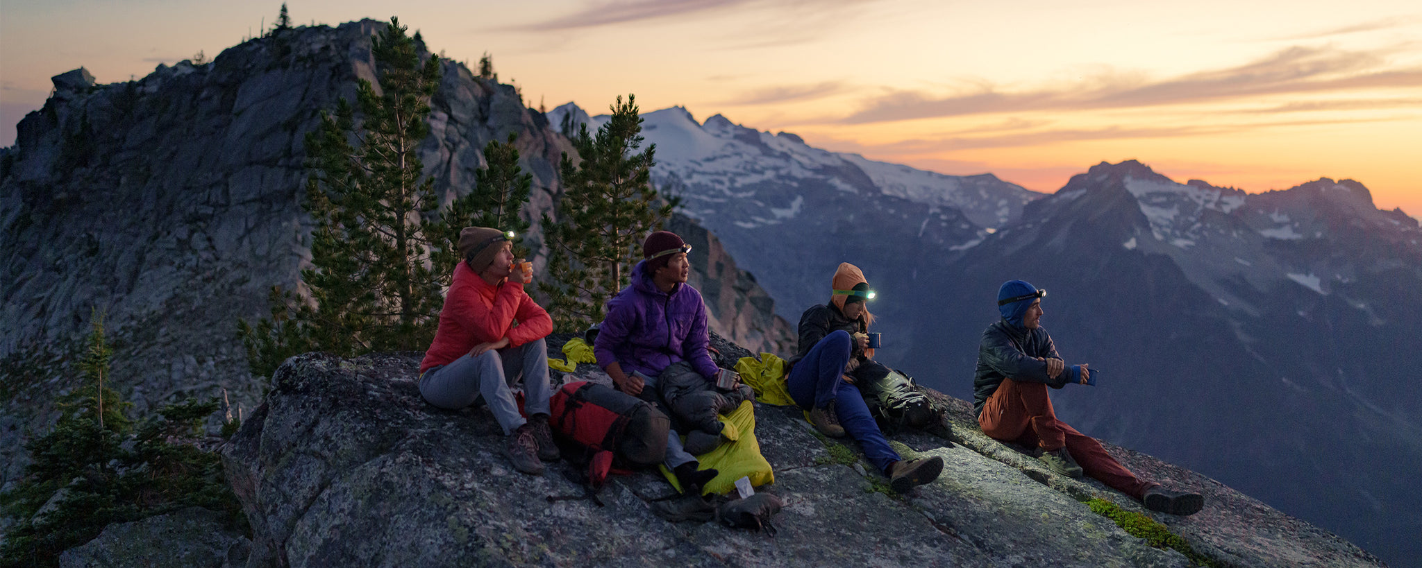 A group of friends watch the sunset while sitting on a rock in the mountains.