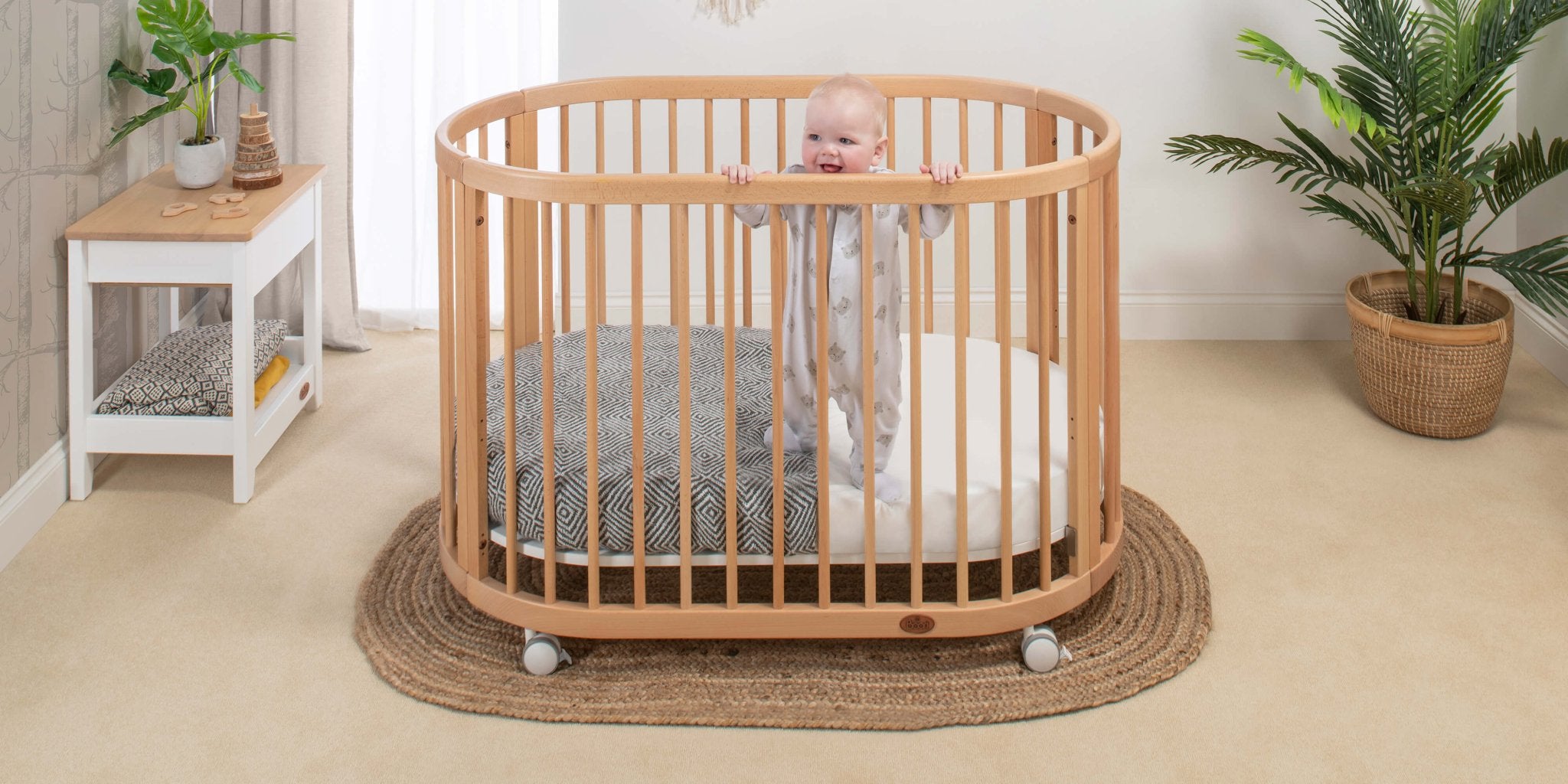 baby in oval cot in light nursery with green plants