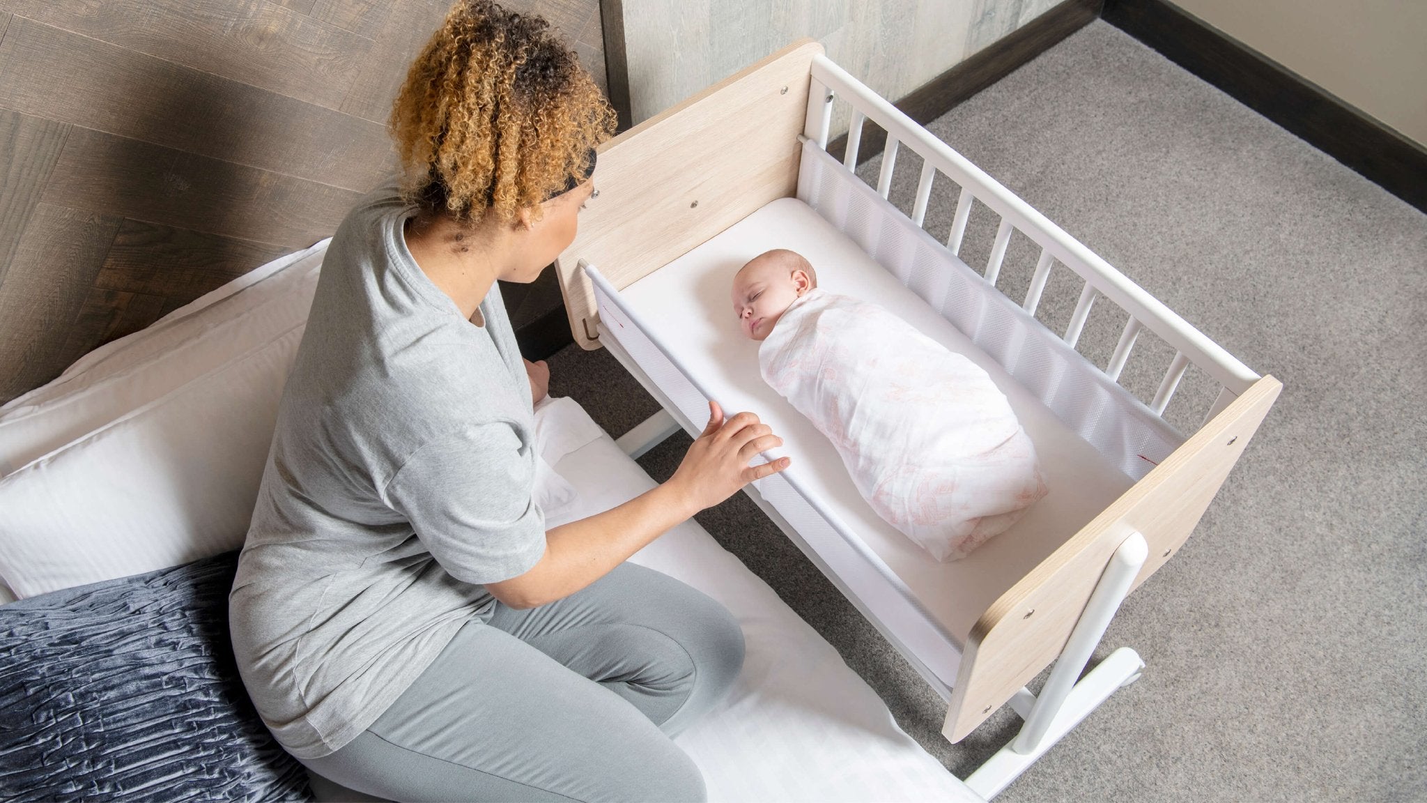 mum rocks swaddled baby in bedside crib