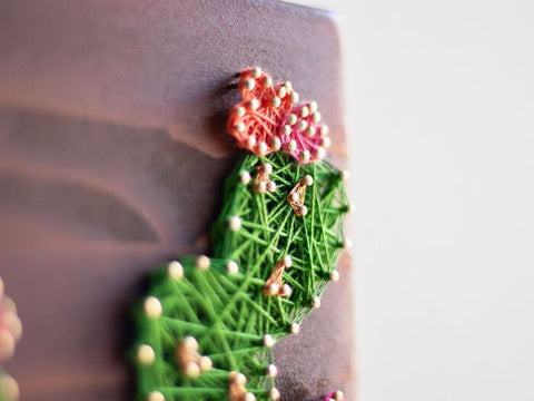 Close-up of a cactus string art with green threads and small pink flowers.