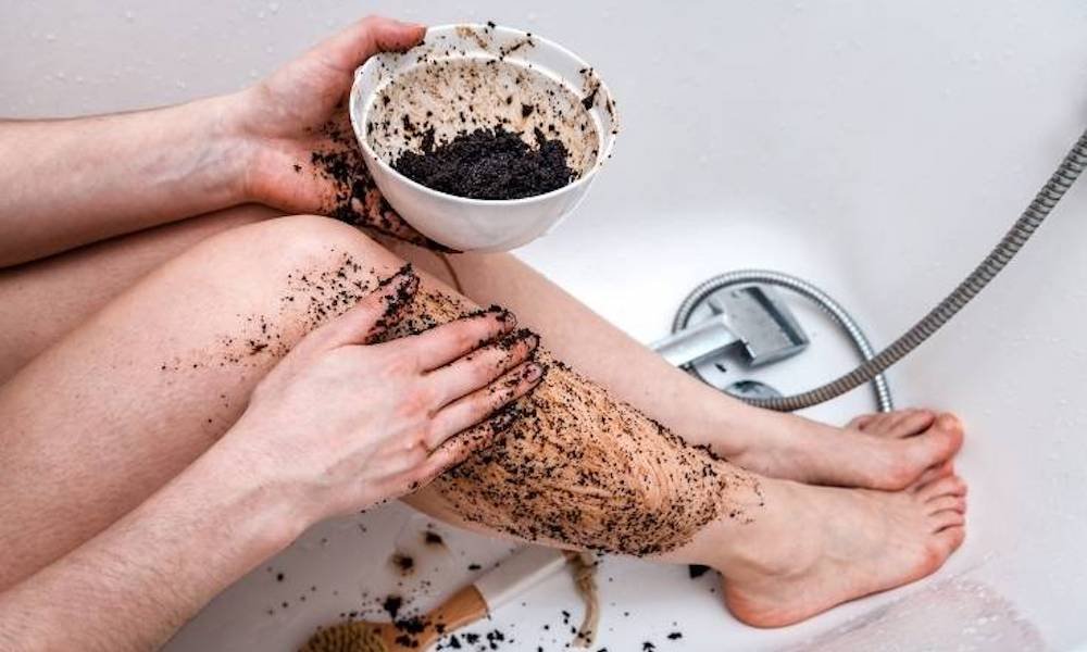 pale legs in white bathtub with hands rubbing coffee grounds exfoliator in