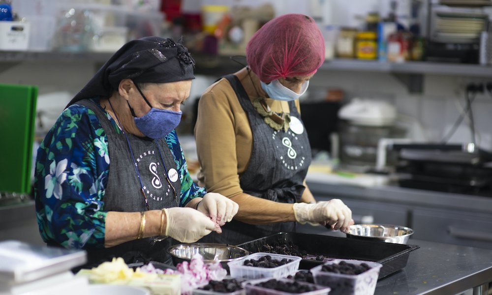 two women wearing gloves and hairnets as they sort blackberries into containers in a small preserving kitchen