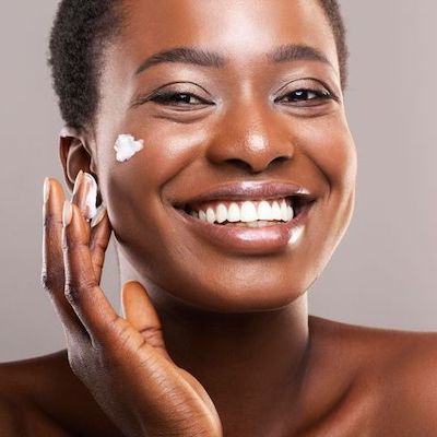 Up close picture of African American woman applying revitalizing cream to her cheek, for our blog post on Revitalizing and Hydrating Facial Skin.