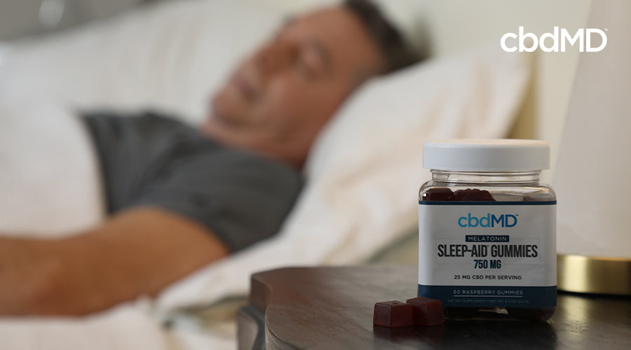 A man sleeping in a bed with a cbdMD melatonin sleep aid gummies 750 mg raspberry container on a nightstand next to him 